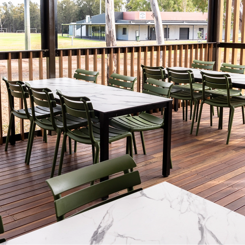 Outdoor dining area on a wooden deck with rectangular tables and green chairs, surrounded by a slatted wooden fence and trees, with a building visible in the background.