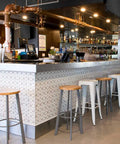 A modern bar interior with a long counter, patterned white and gray tile, multiple white bar stools with wood tops, copper piping overhead, and shelves of bottles in the background.
