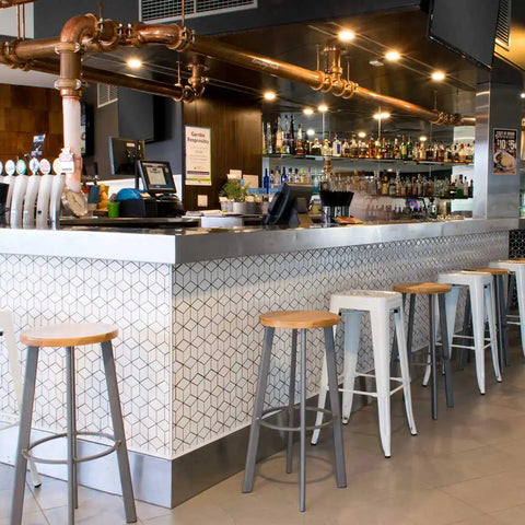 A modern bar interior with a long counter, patterned white and gray tile, multiple white bar stools with wood tops, copper piping overhead, and shelves of bottles in the background.